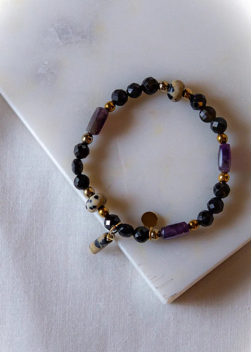 Królewska Ochrona Bransoletka Z Turmalinu I Ametystu: ręcznie wykonana bransoletka z kamieni czarnego turmalinu, ametystu, z metalicznymi złotymi koralikami, leży na białej abstrakcyjnej powierzchni.