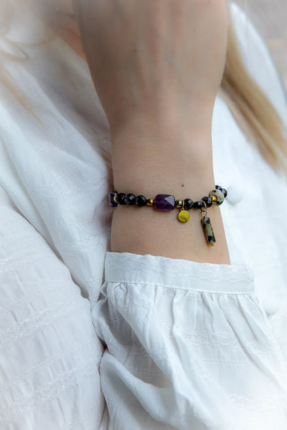 Królewska Ochrona Bransoletka Z Turmalinu I Ametystu Biżuteria: turkusowy kwadratowy ametyst otoczony czarnymi kamieniami, złotymi koralikami i wiszącymi zawieszkami, na nadgarstku w białej, haftowanej bluzce.