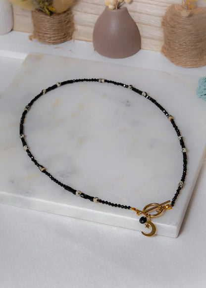 Księżycowy Talizman Naszyjnik Czarny Turmalin na białym tle, złoty zapięcie z uroczym księżycem i gwiazdą.