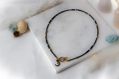 Księżycowy Talizman Naszyjnik Czarny Turmalin złożony z czarnych i białych koralików, ozdobiony złotym zapięciem i zawieszką w kształcie księżyca.