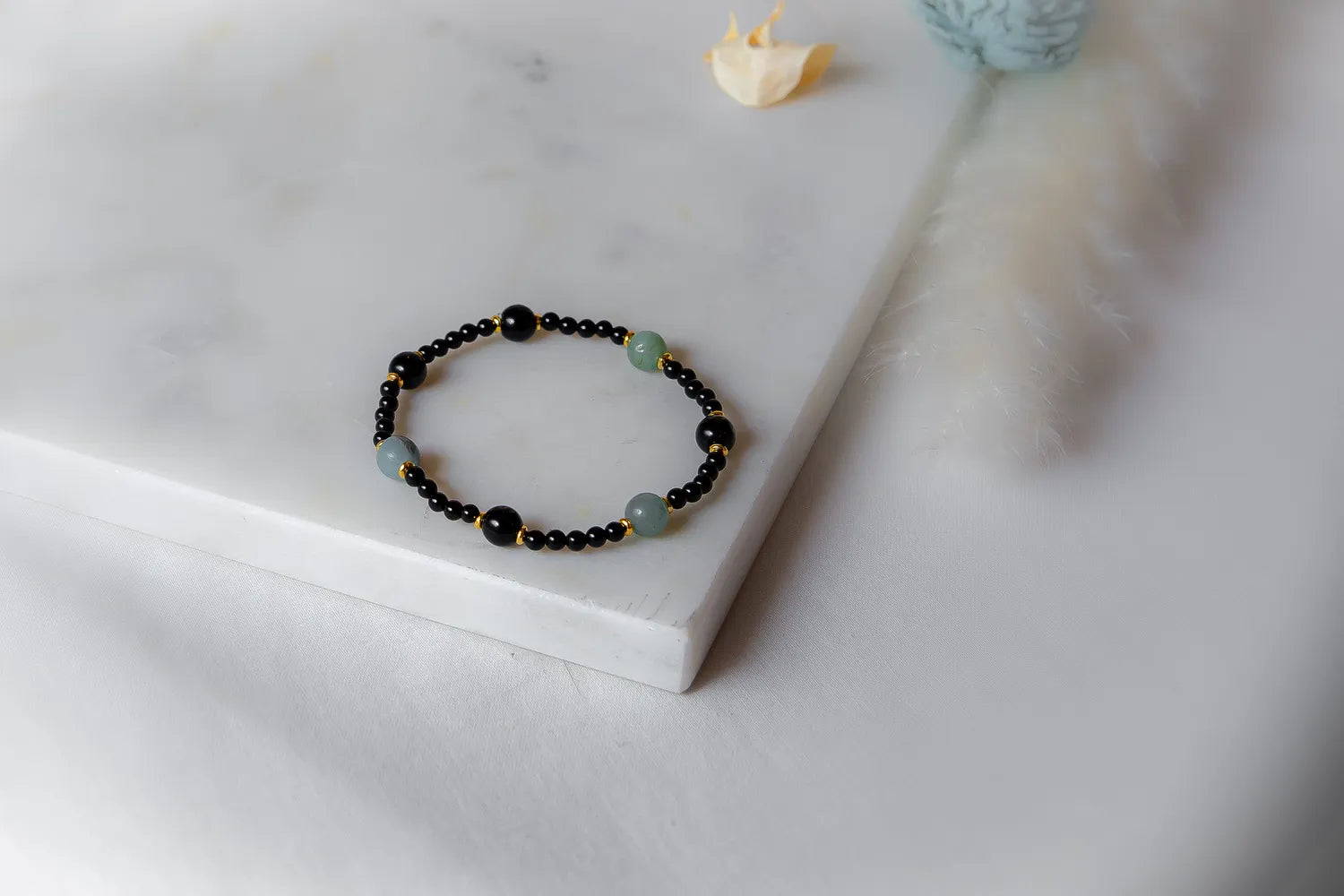 Lodowa Elegancja Bransoletka Agat Opal z czarnymi i jasnozielonymi kamieniami na marmurowym tle z piórkiem.