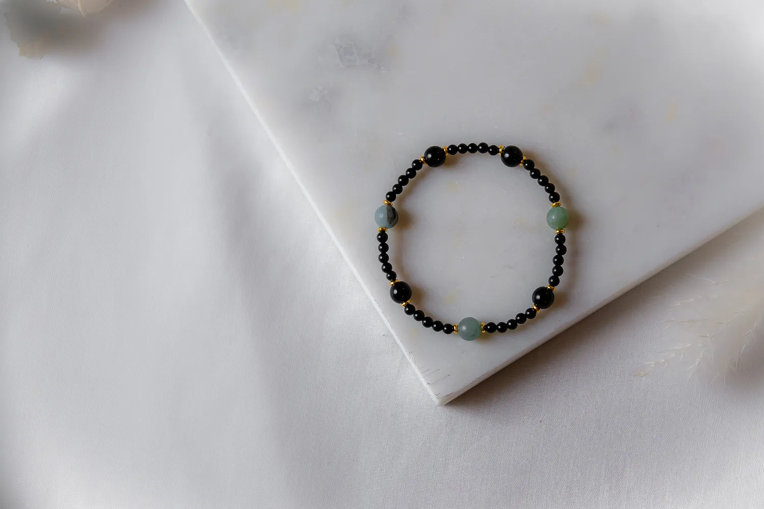 Lodowa Elegancja Bransoletka Agat Opal: czarna, drobna ośmiorniczka z pojedynczymi, większymi kamieniami agatu w odcieniach szarości i zieleni, przeplatana złotymi koralikami, ułożona na białej, marmurowej powierzchni.