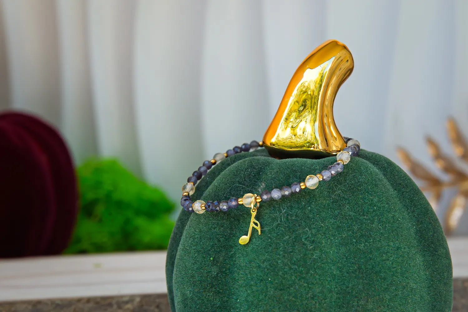 MELODIA MAGII - BRANSOLETKA Z LABRADORYTU I CYTRYNU, prezentowana na zielonym aksamitnym podstawce z złotym akcentem w tle. Bransoletka ozdobiona jest koralikami z labradorytu i cytrynu oraz złotym elementem w kształcie nuty muzycznej.