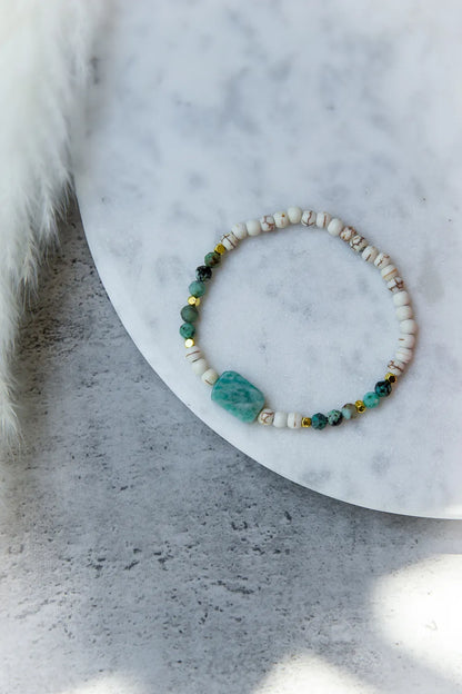 Morska Bryza Bransoletka Kamienie – Płasko ułożona, z akwamarynowym kamieniem centralnym i turkusowymi kuleczkami na białym tle.