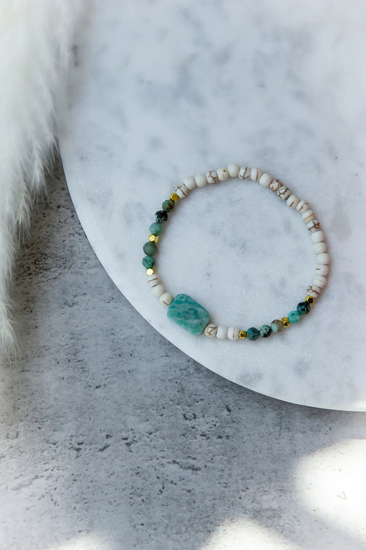Morska Bryza Bransoletka Kamienie – Płasko ułożona, z akwamarynowym kamieniem centralnym i turkusowymi kuleczkami na białym tle.