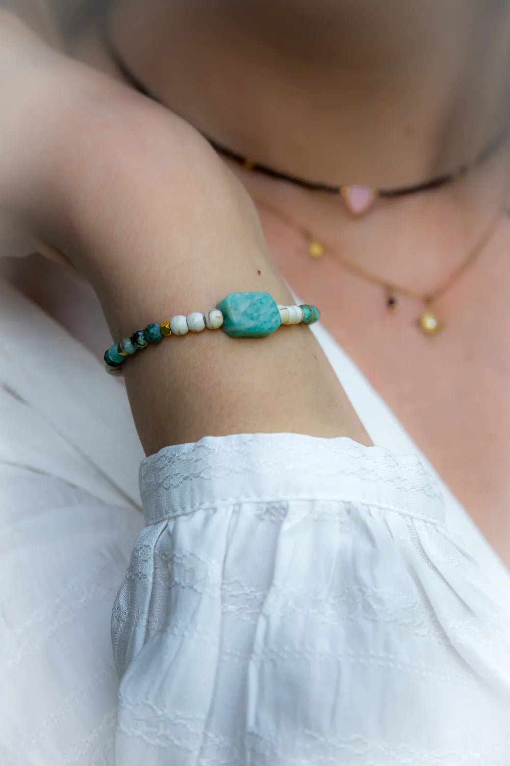 Morska Bryza Bransoletka Z Kamieniami na damskiej ręce z turkusowymi, białymi i złotymi koralikami, na bladej skórze, w tle inne naszyjniki.