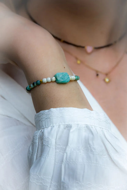 Morska Bryza Bransoletka Z Kamieniami na damskiej ręce z turkusowymi, białymi i złotymi koralikami, na bladej skórze, w tle inne naszyjniki.