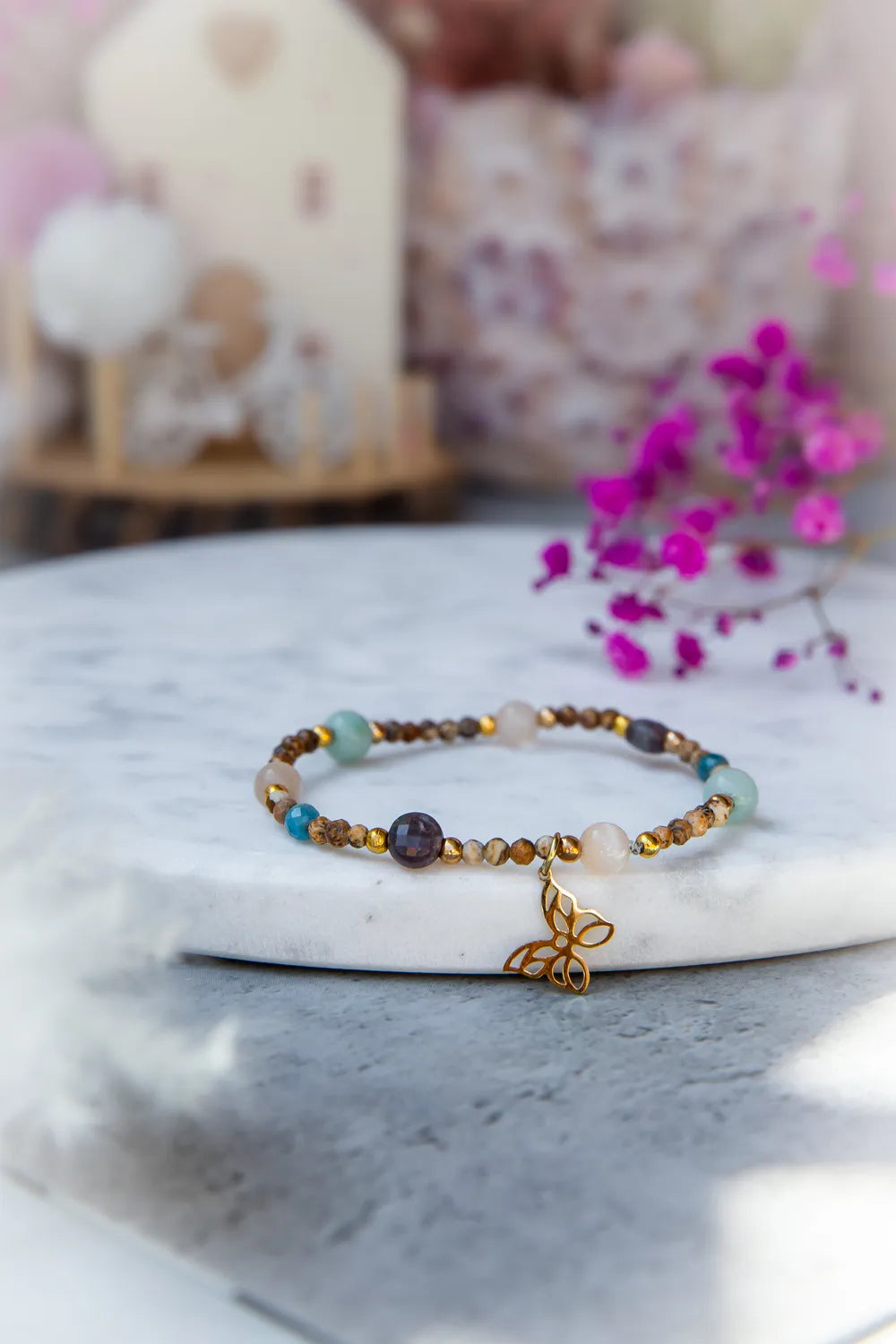 Motyli Taniec Bransoletka Z Kamieniami, z koralików w odcieniach brązu, zieleni i różu, ozdobiona złotym motylkiem, na marmurowym tle z fuksją.