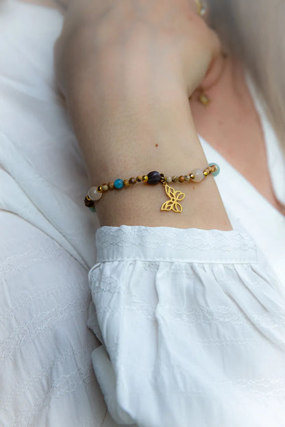 Motyli Taniec Bransoletka Z Kamieniami na nadgarstku, zdobiona złotym motylem i wielobarwnymi kamieniami, na tle białej, zwiewnej tkaniny.