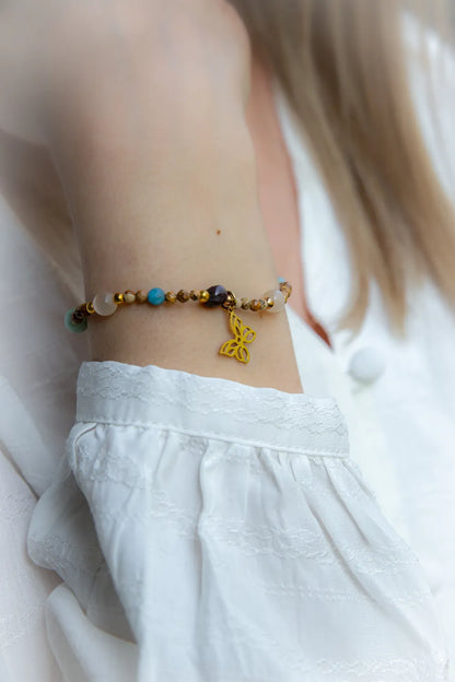 Motyli Taniec Bransoletka Z Kamieniami: złoty motyl zawieszka na nadgarstku, z koralikami: niebieskimi, białymi, brązowo-złotymi i fioletowymi. W tle jasna, delikatna tkanina.