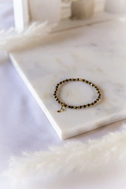 Muzyczna Nutka Bransoletka Dla Fana Muzyki Elegancka na marmurowej podstawce, z czarnymi i złotymi kamieniami oraz złotą zawieszką w kształcie nutki.