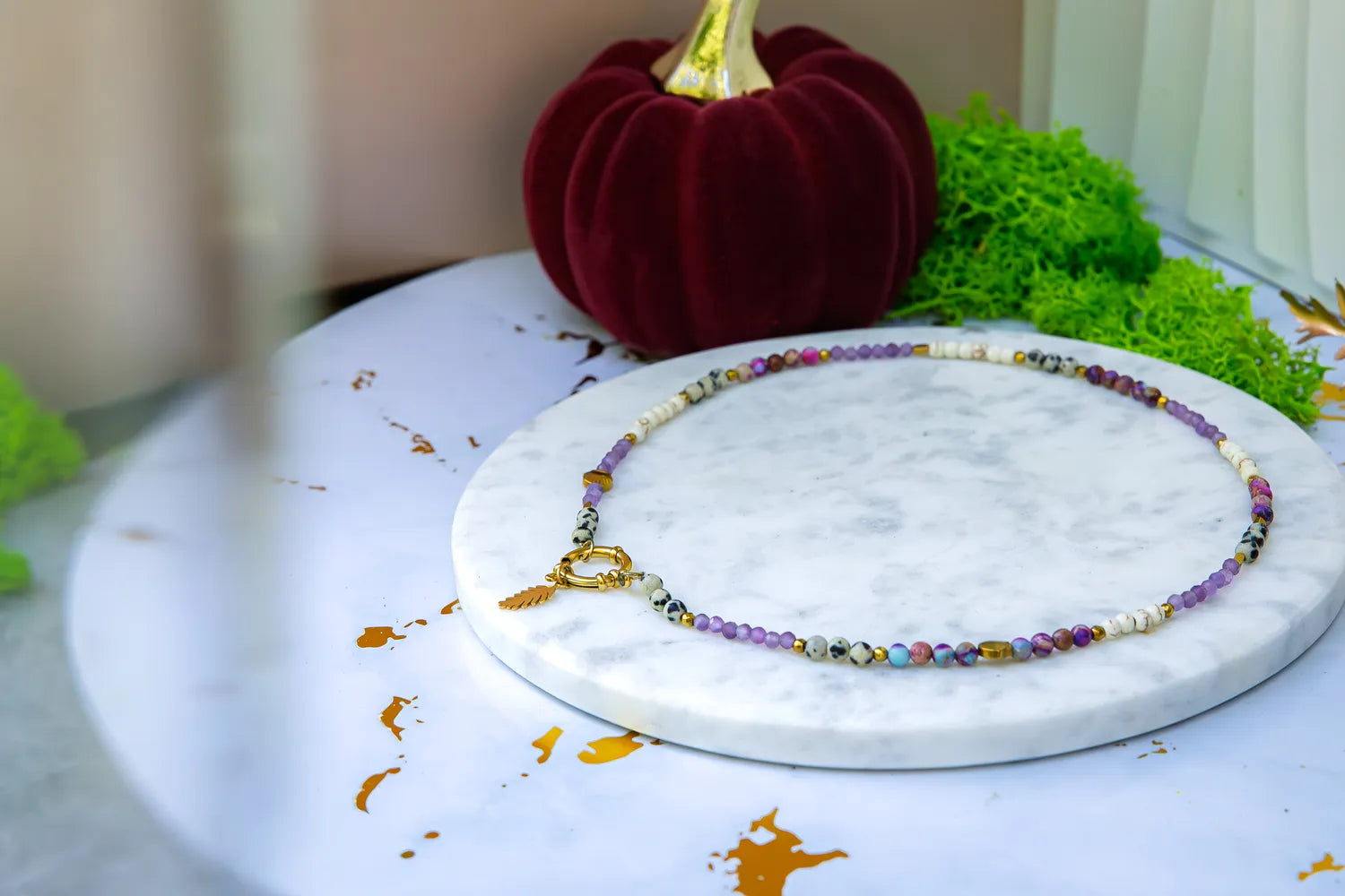 PEŁNIA HARMONII - NASZYJNIK Z AMETYSTU, HOWLITU I JASPISU DALMATYŃSKIEGO. Naszyjnik z kolorowymi kamieniami układa się wokół białej marmurowej podstawki, z czerwoną dynią i zielonym mchem w tle.