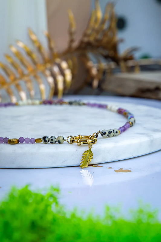 PEŁNIA HARMONII - NASZYJNIK Z AMETYSTU, HOWLITU I JASPISU DALMATYŃSKIEGO. Naszyjnik z kolorowymi koralikami ametystu, howlitu i jaspisu dalmatyńskiego, ułożony na białym marmurowym tle z złotym liściem wisiorem. Tło ozdobione złotą gałązką.