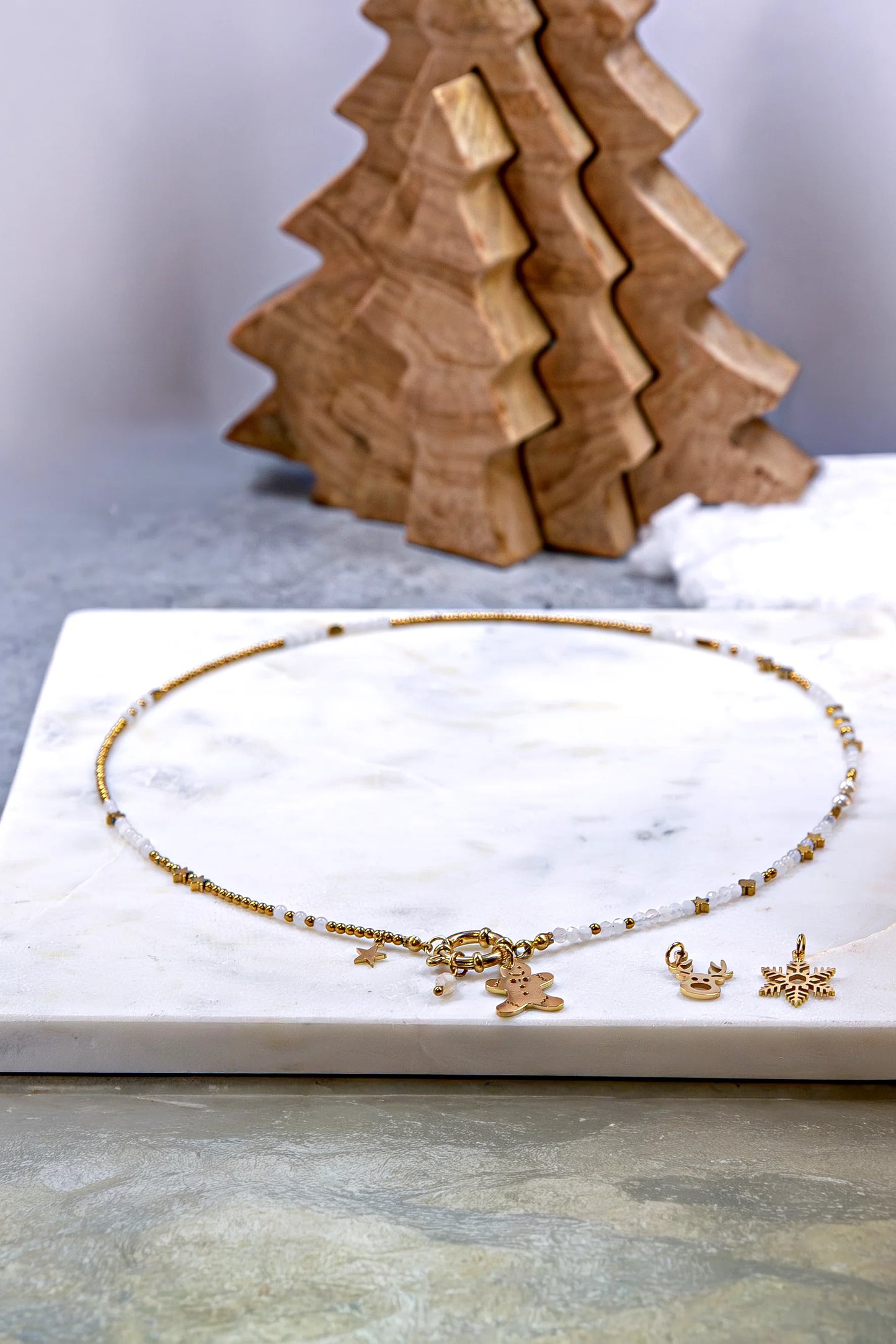 Naszyjnik z główną zawieszką w kształcie ludzika z piernika. Obok leżą dodatkowe, złote charmsy do wyboru: głowa renifera i śnieżynka. Biżuteria celebrująca indywidualność.