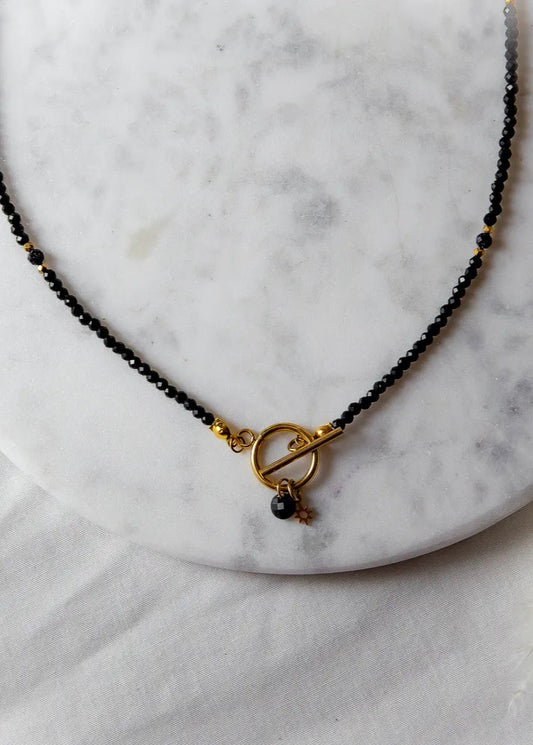 Noc Kairu Naszyjnik Czarny Kamienie z złotym zapięciem toggle i zawieszką gwiazdką, na marmurowej podstawie z białym futerkiem w tle.