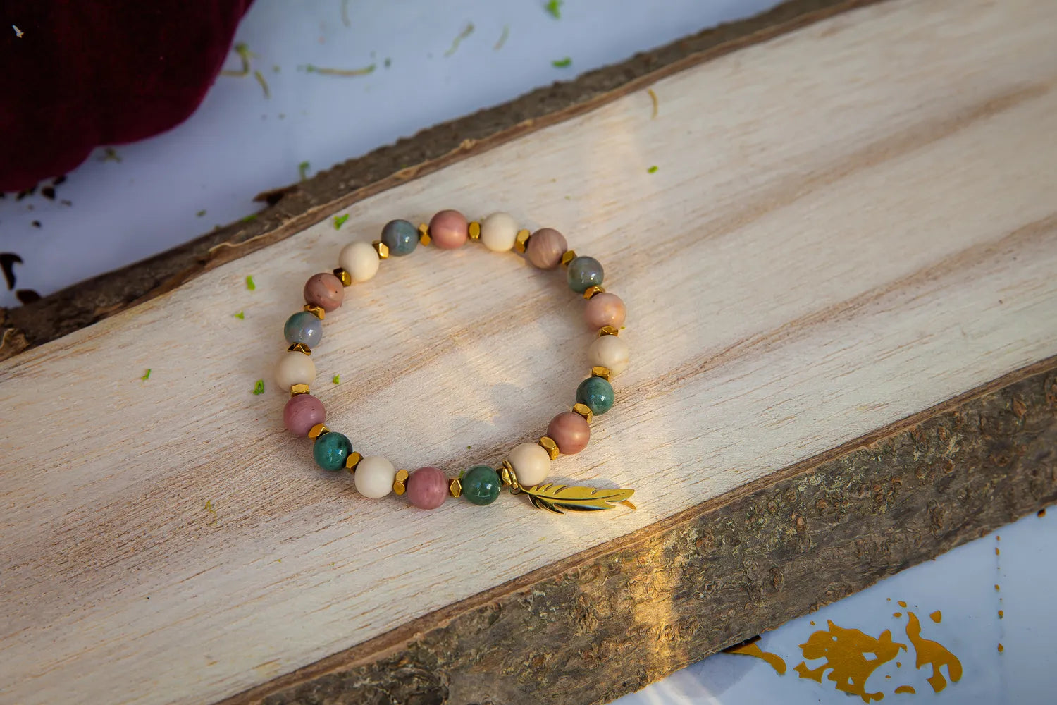 PAMIĘĆ ZIEMI - BRANSOLETKA Z KAMIENIA FOSYLNEGO, RODONITU I AGATU. Bransoletka z kolorowymi koralikami z kamienia fosylnego, rodonitu i agatu, ułożona na drewnianym tle z dodatkiem złotej piórka.