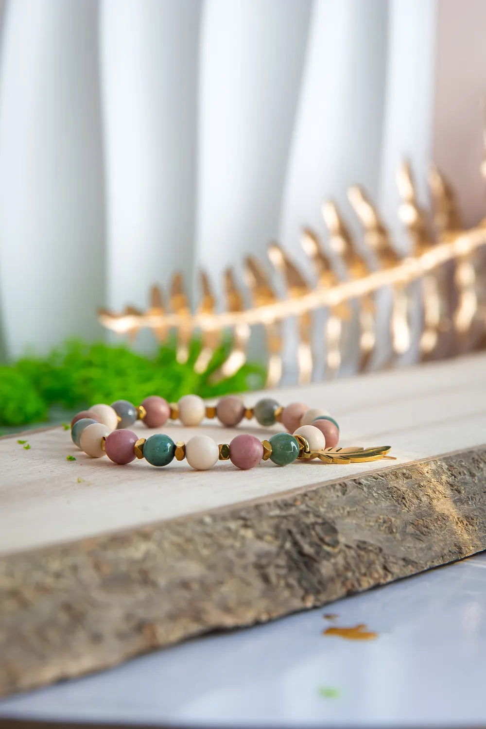 PAMIĘĆ ZIEMI - BRANSOLETKA Z KAMIENIA FOSYLNEGO, RODONITU I AGATU. Bransoletka z kolorowymi koralikami z kamienia fosylnego, rodonitu i agatu, ułożona na drewnianym tle z zielonymi liśćmi i złotym elementem w tle.