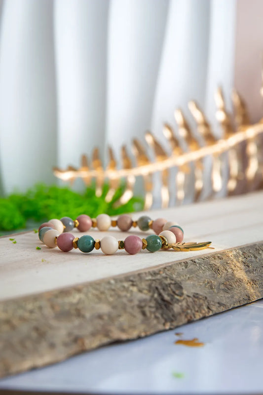 PAMIĘĆ ZIEMI - BRANSOLETKA Z KAMIENIA FOSYLNEGO, RODONITU I AGATU. Bransoletka z kolorowymi koralikami z kamienia fosylnego, rodonitu i agatu, ułożona na drewnianym tle z zielonymi liśćmi i złotym elementem w tle.