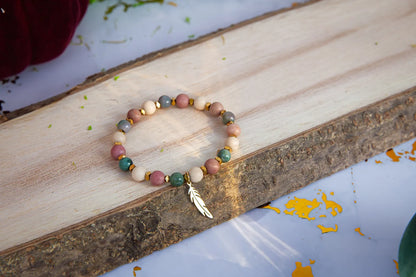Bransoletka 'PAMIĘĆ ZIEMI' z kamieniem fosylnym, rodonitem i agatem, ułożona na drewnianym tle z dodatkiem liścia. Biżuteria ręcznie robiona z kolorowymi koralikami.