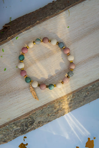 Bransoletka 'PAMIĘĆ ZIEMI' z kamieniem fosylnym, rodonitem i agatem, ułożona na drewnianym tle z dodatkiem złotego elementu w kształcie liścia.