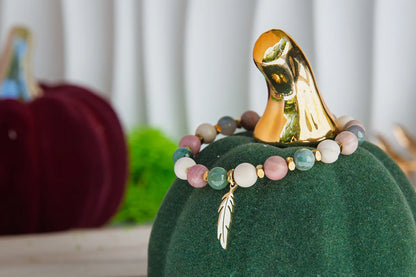 PAMIĘĆ ZIEMI - BRANSOLETKA Z KAMIENIA FOSYLNEGO, RODONITU I AGATU. Bransoletka z kolorowymi kamieniami i złotym liściem zawieszonym na zielonym dyniowym tle.