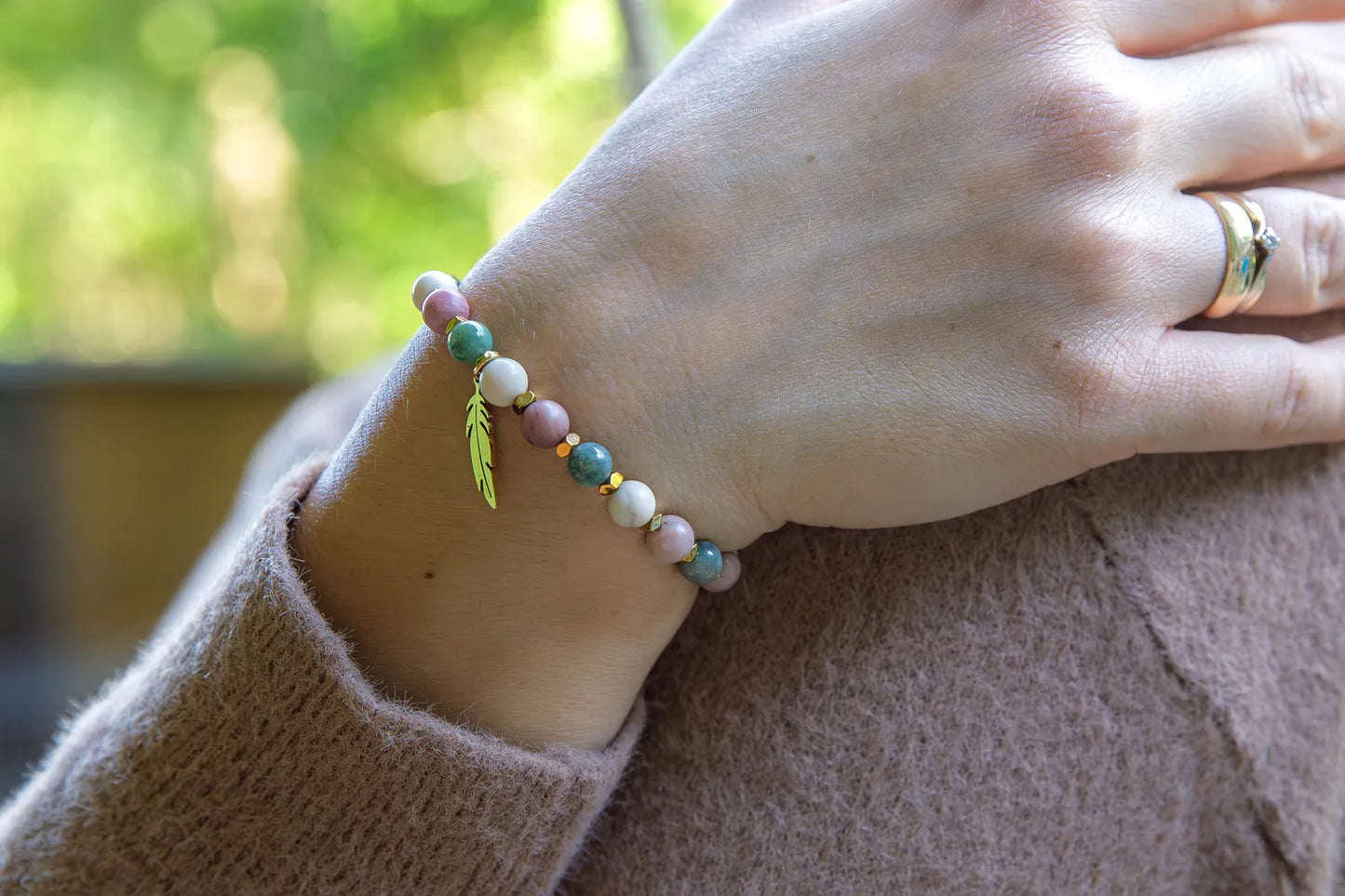 PAMIĘĆ ZIEMI - BRANSOLETKA Z KAMIENIA FOSYLNEGO, RODONITU I AGATU. Bransoletka z kolorowymi koralikami z kamienia fosylnego, rodonitu i agatu, ozdobiona złotym liściem. Tło zielone, ułożenie na nadgarstku.