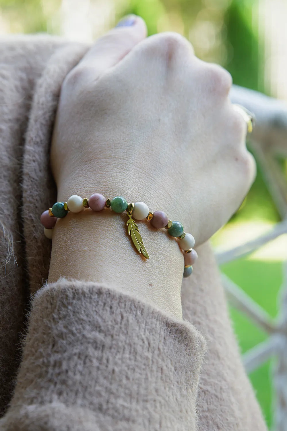Bransoletka 'PAMIĘĆ ZIEMI' z kamieniem fosylnym, rodonitem i agatem na nadgarstku, ozdobiona złotym liściem. Tło zielone, ułożenie naturalne.