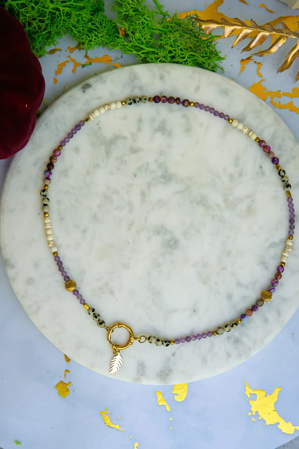 PEŁNIA HARMONII - NASZYJNIK Z AMETYSTU, HOWLITU I JASPISU DALMATYŃSKIEGO. Naszyjnik z kolorowymi kamieniami ametystu, howlitu i jaspisu dalmatyńskiego, ułożony na białym marmurowym tle. W tle zielone i złote dekoracje.