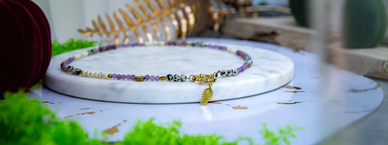 PEŁNIA HARMONII - NASZYJNIK Z AMETYSTU, HOWLITU I JASPISU DALMATYŃSKIEGO. Naszyjnik z kolorowymi kamieniami ametystu, howlitu i jaspisu dalmatyńskiego, ułożony na białym marmurowym podstawce z zielonymi dodatkami w tle.