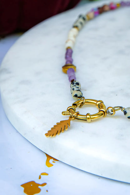 PEŁNIA HARMONII - NASZYJNIK Z AMETYSTU, HOWLITU I JASPISU DALMATYŃSKIEGO. Naszyjnik z kolorowymi kamieniami, w tym ametystem, howlitem i jaspisem dalmatyńskim, na białym marmurowym tle. Złoty element z liściem.