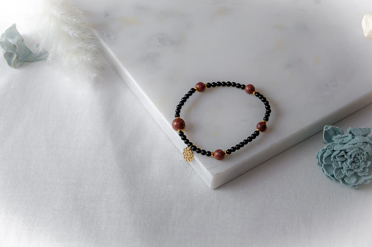 Pustynna Elegancja Bransoletka Piasku Pustyni: czarne koraliki z błyszczącymi, brązowymi kamieniami piaskowymi i złotą zawieszką na białym tle.