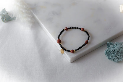 Pustynna Elegancja Bransoletka Piasku Pustyni: czarne koraliki z błyszczącymi, brązowymi kamieniami piaskowymi i złotą zawieszką na białym tle.