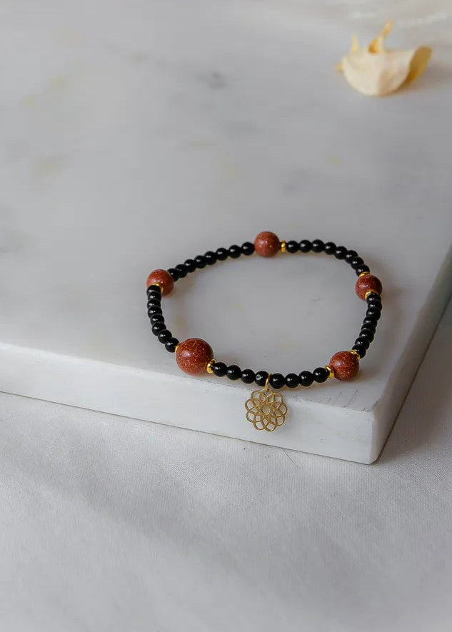 Pustynna Elegancja Bransoletka Piasku Pustyni: czarne koraliki z ośmioma błyszczącymi, pomarańczowymi kamieniami pustyni i złotym zawieszką w kształcie kwiatu.