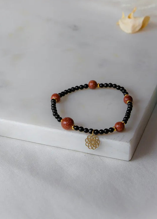 Pustynna Elegancja Bransoletka Piasku Pustyni: czarne koraliki z ośmioma błyszczącymi, pomarańczowymi kamieniami pustyni i złotym zawieszką w kształcie kwiatu.