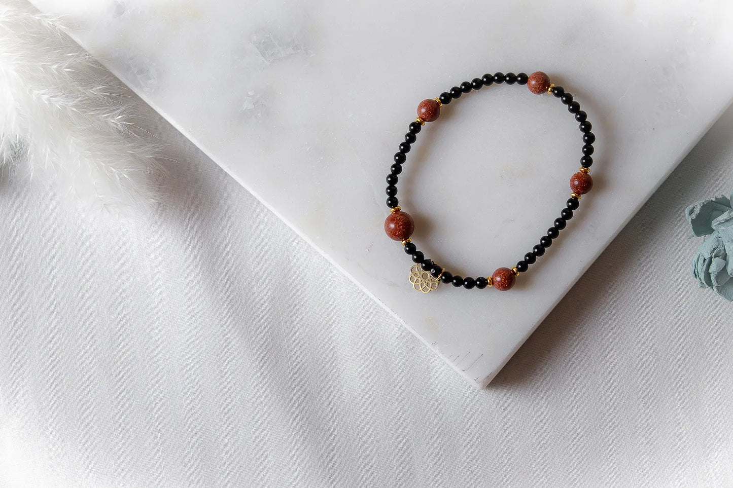 Pustynna Elegancja Bransoletka Piasku Pustyni z czarnymi i brązowymi kamieniami oraz złotym elementem na marmurowej powierzchni.