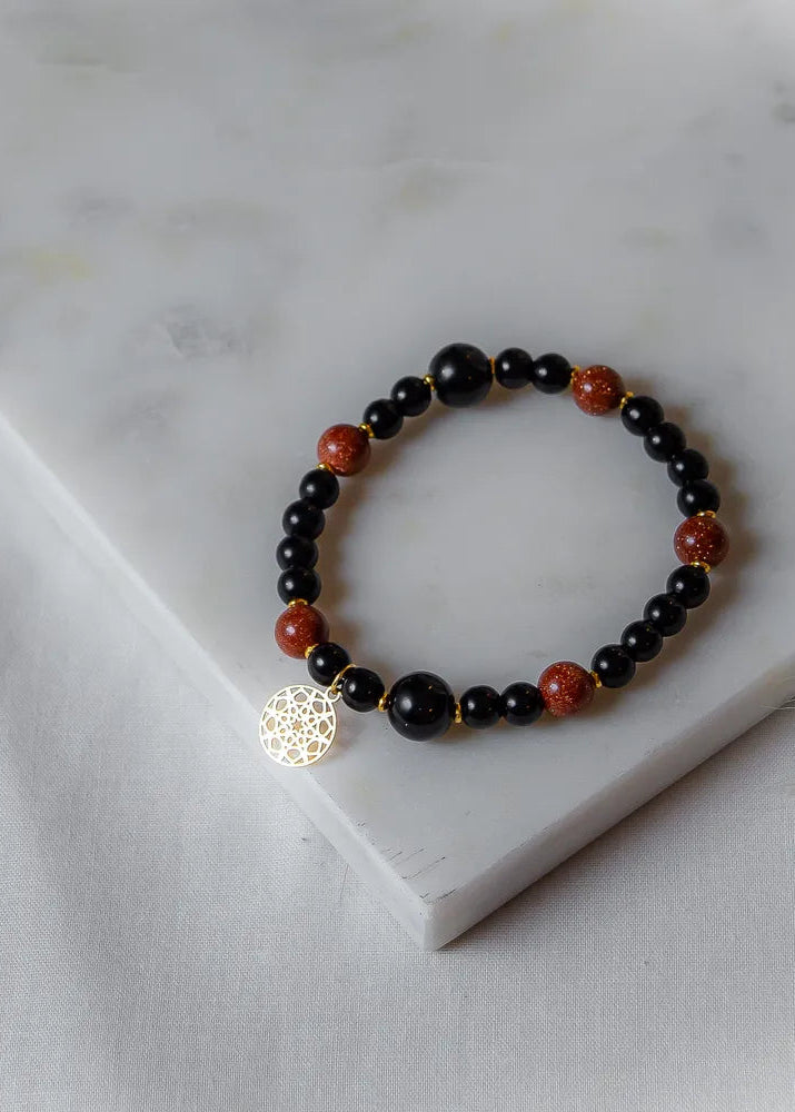 Pustynna Moc Bransoletka Xl Piasku Pustyni: czarne i złociste kamienie na białym tle, ozdobiona złotą zawieszką.