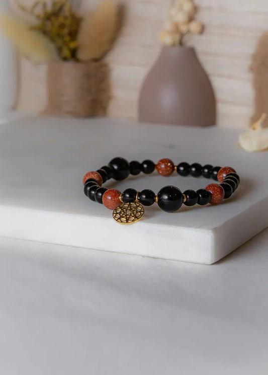 Pustynna Moc Bransoletka Xl Piasku Pustyni Czarny Onyks - z czarnymi i błyszczącymi kamieniami, ozdobiona złotym medalionem, na marmurowej platformie.