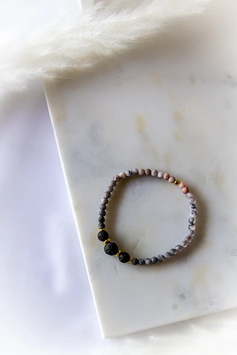 Różane Ognisko Bransoletka Rodonit Lawa ze złotymi przekładkami, o matowym wykończeniu. Ułożona na białym marmurze z delikatnym pióropuszem.