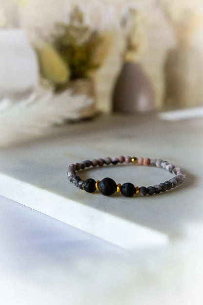 Różane Ognisko Bransoletka Z Rodonitu I Lawy: delikatne kamienie rodonitu, czarne kulki lawy i złote przekładki na jasnym marmurkowym tle.