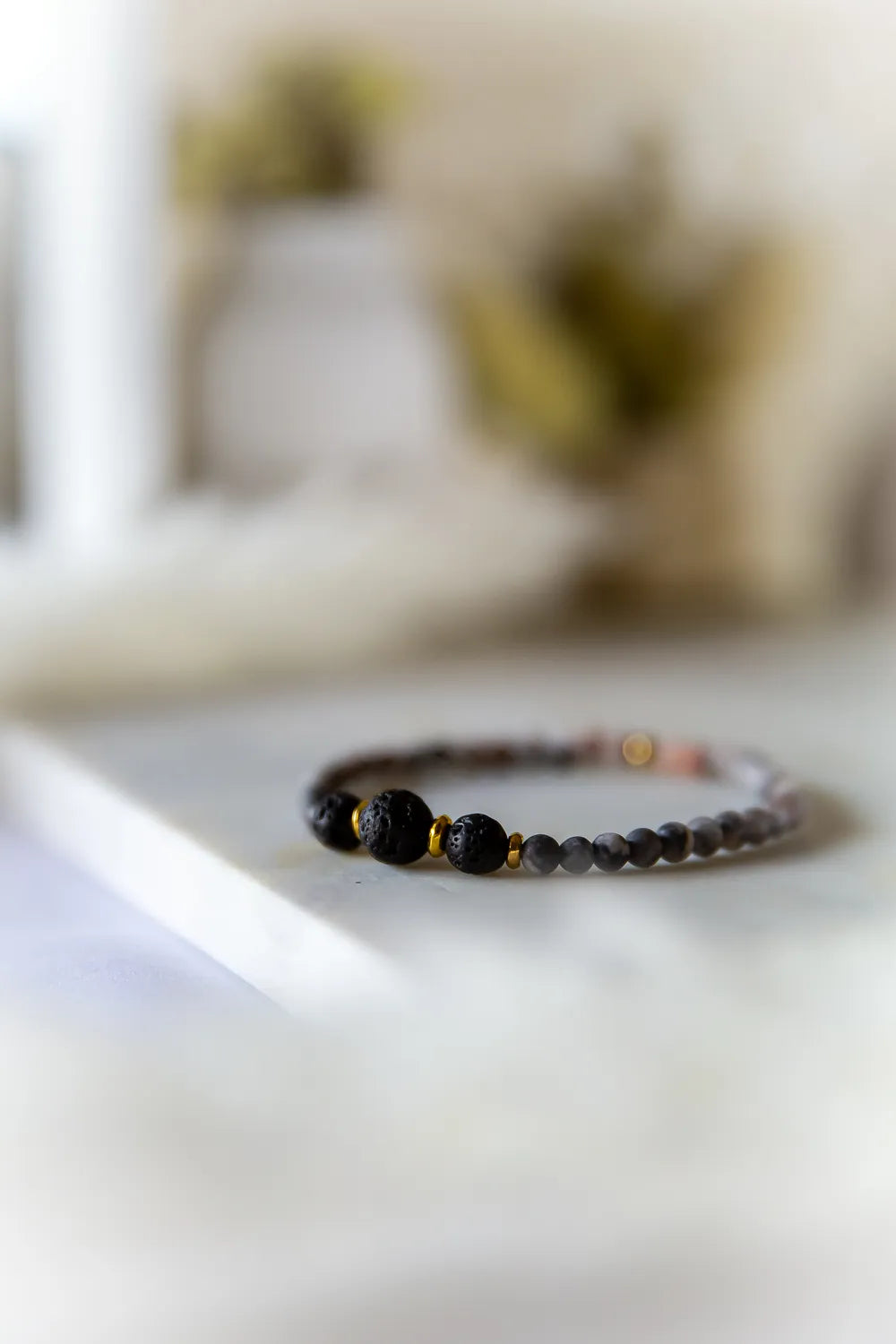 Różane Ognisko Bransoletka Z Rodonitu I Lawy: zbliżenie na bransoletkę z czarnymi kamieniami lawy i drobnymi, szarymi kamieniami rodonitu, ozdobiona złotymi przekładkami.