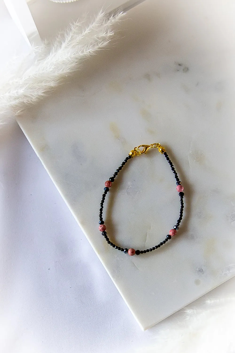 Różowa Ochrona Bransoletka Z Turmalinu I Jaspisu na marmurowej powierzchni z delikatną piórkową ozdobą. Kilka różowych kamieni z turmalinu i jaspisu przeplatane czarnymi, błyszczącymi koralikami.