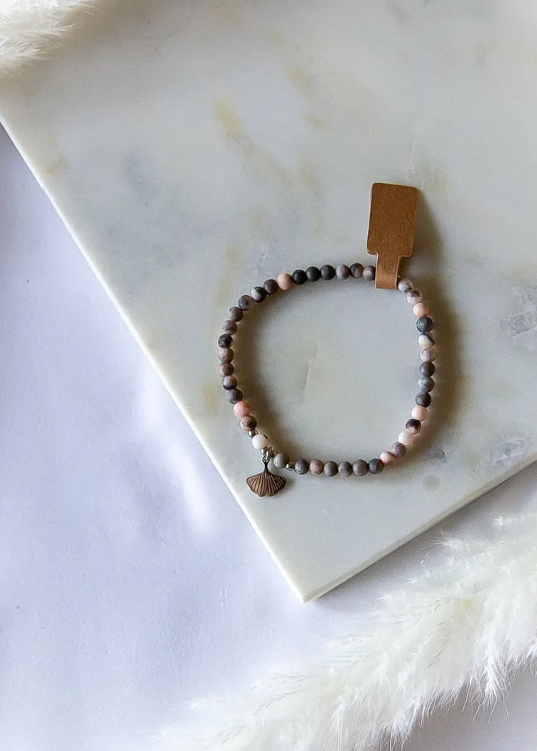 Różowy Zen Bransoletka Rodonit Zawieszka z metalową zawieszką w kształcie liścia miłorzębu na tle marmurowej płyty i naturalnych ozdób.