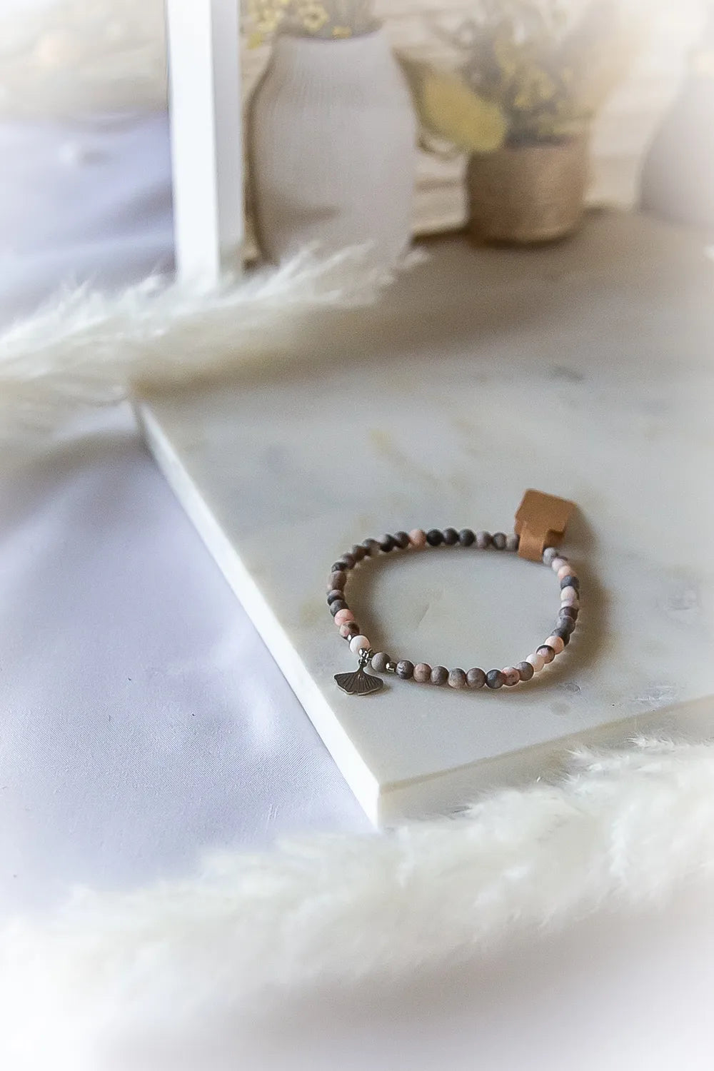 Różowy Zen Bransoletka Rodonit Zawieszka z dyskretną zawieszką w kształcie liścia miłorzębu na marmurowej powierzchni, z dodatkiem ozdobnych traw na tle.