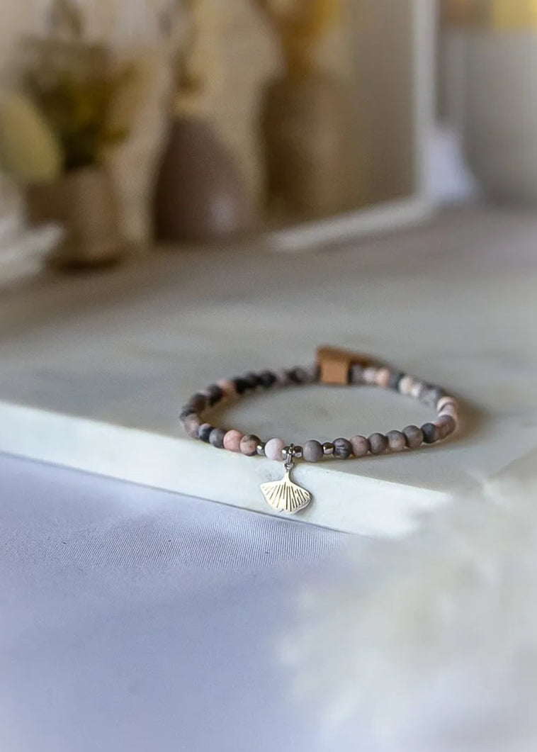 Różowy Zen Bransoletka Z Rodonitu ze srebrną zawieszką w kształcie liścia miłorzębu, na kamiennym tle z subtelnym rozmyciem.