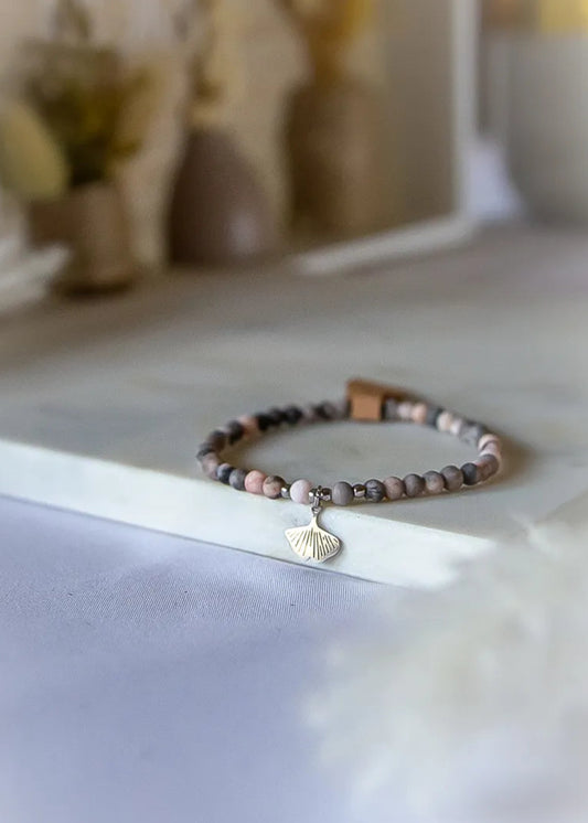 Różowy Zen Bransoletka Z Rodonitu ze srebrną zawieszką w kształcie liścia miłorzębu, na kamiennym tle z subtelnym rozmyciem.