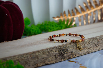 Bransoletka 'SERCE JESIENI' z kamieniami karneolu, granatu i cytrynu, ułożona na drewnianym tle z zielonymi liśćmi w tle.