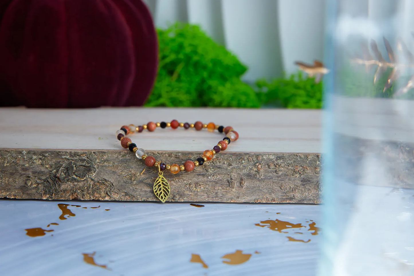 Bransoletka 'SERCE JESIENI' z karneolu, granatu i cytrynu, ozdobiona złotym liściem, leży na drewnianym bloku z zielonym tłem i pomarańczową dynią w tle.
