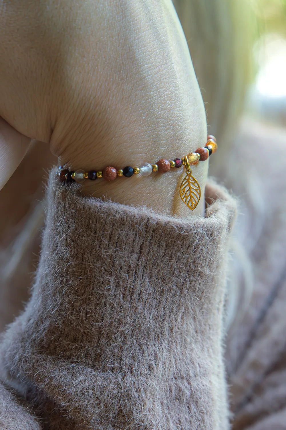 Bransoletka 'SERCE JESIENI' z karneolu, granatu i cytrynu, ozdobiona złotym liściem, na tle delikatnej, beżowej tkaniny.