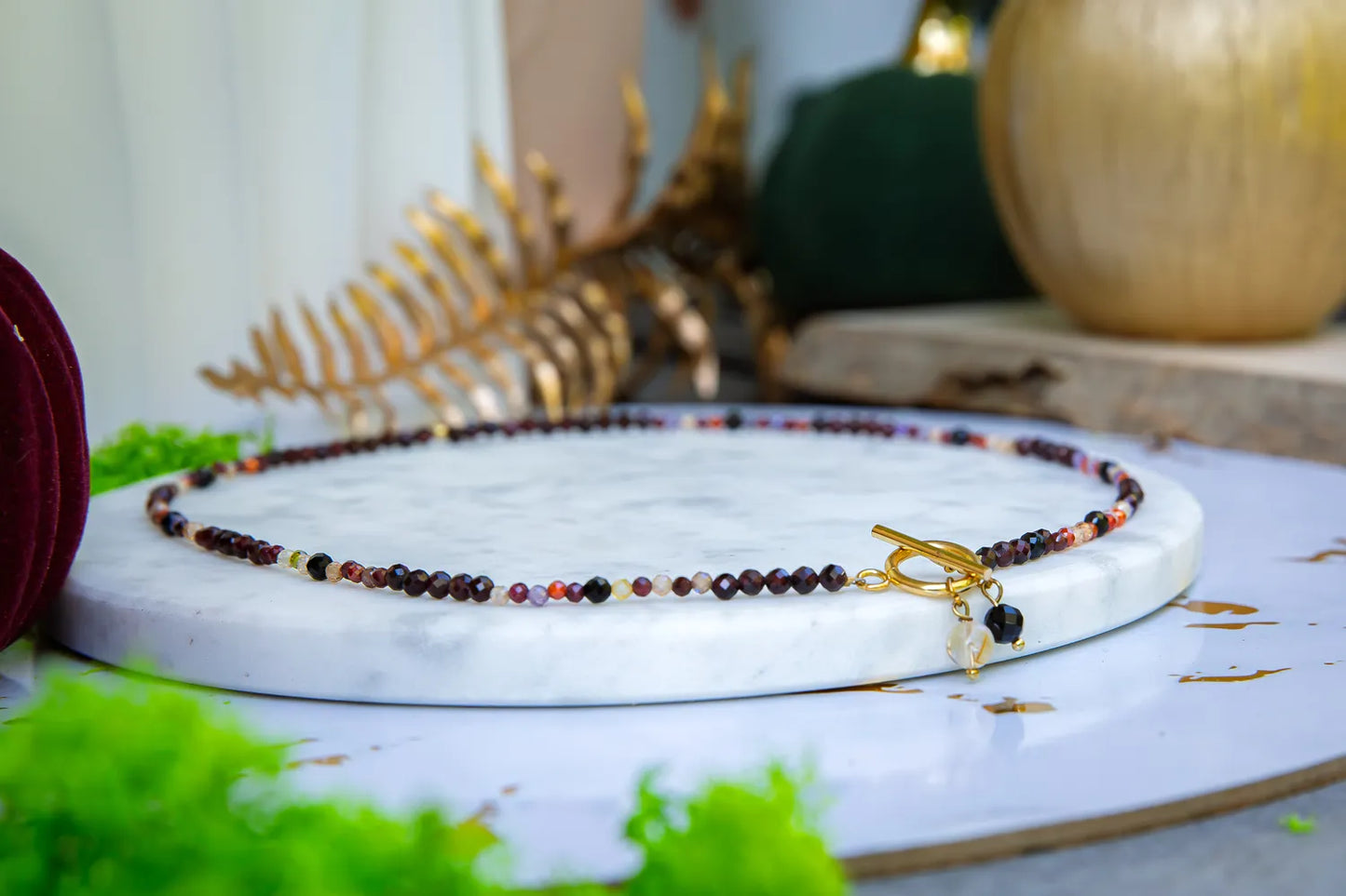 SERCE KRÓLOWEJ - NASZYJNIK Z GRANATU, SPINELU I CYTRYNU. Naszyjnik z kolorowymi kamieniami na białym marmurowym tle, z zielonymi liśćmi i złotą ozdobą w tle.