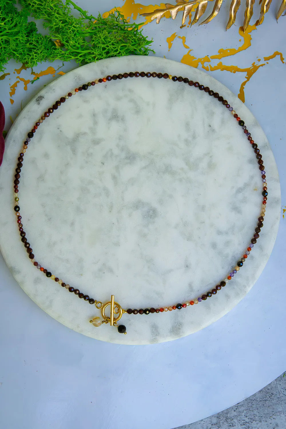 SERCE KRÓLOWEJ - NASZYJNIK Z GRANATU, SPINELU I CYTRYNU, elegancki naszyjnik z kolorowymi kamieniami ułożony na marmurowym tle, z zielonymi i złotymi dodatkami w tle.